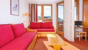 Una habitación con sofá rojo, un banco de madera, un escritorio con computadora y una ventana con vistas a montañas.