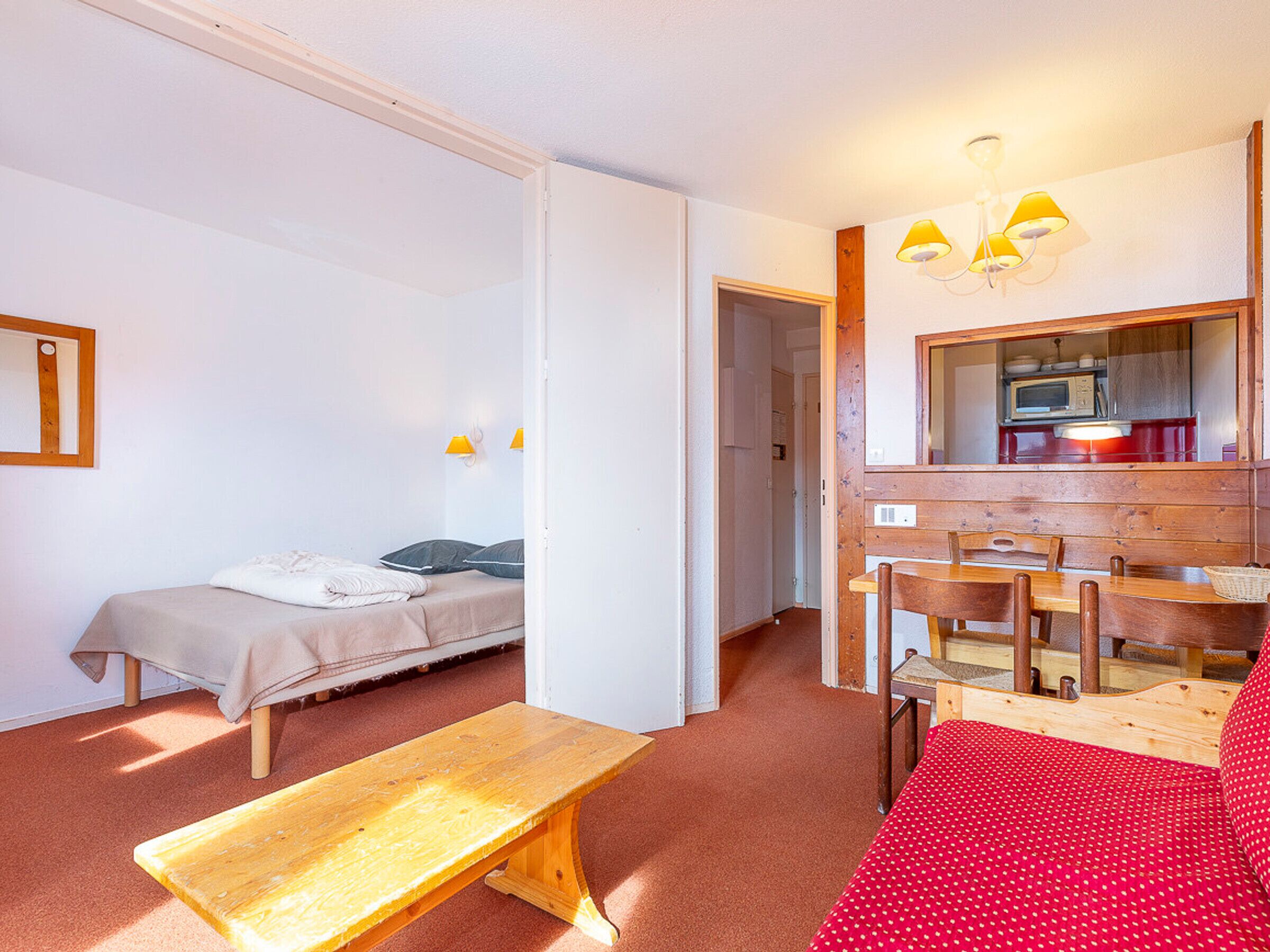 A room with a bed, a bench, a dining table with chairs, a mirror, and a kitchenette.