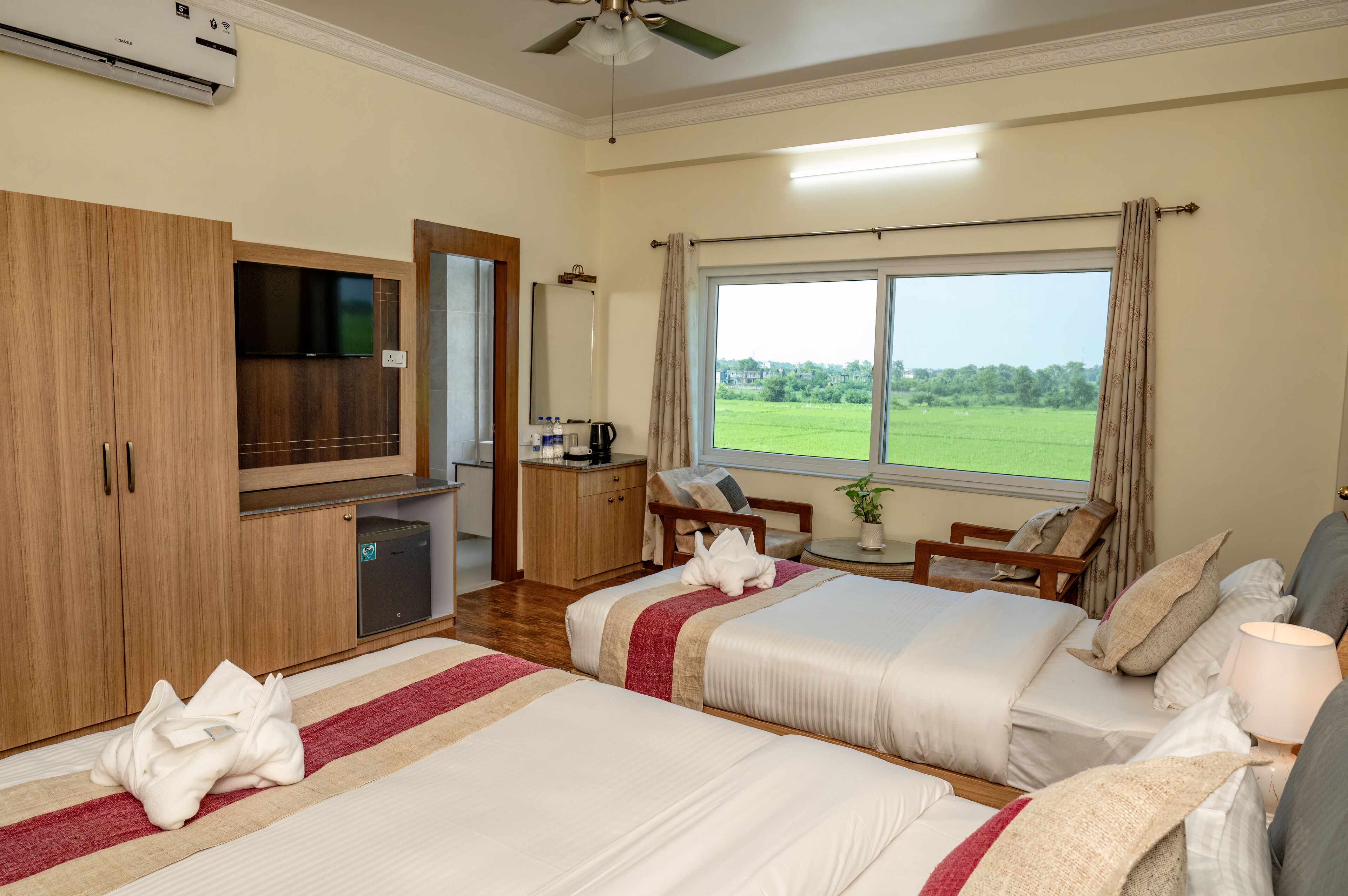A hotel room with two beds, a TV, a desk, a chair, and a window with a view of a green field.