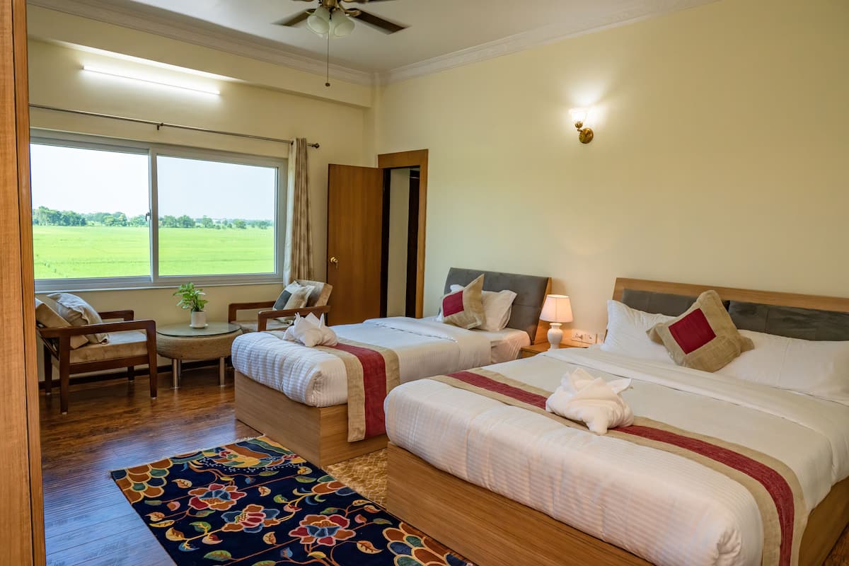 A hotel room with two beds, a small table with a plant, a chair, and a view of a green field through a large window.