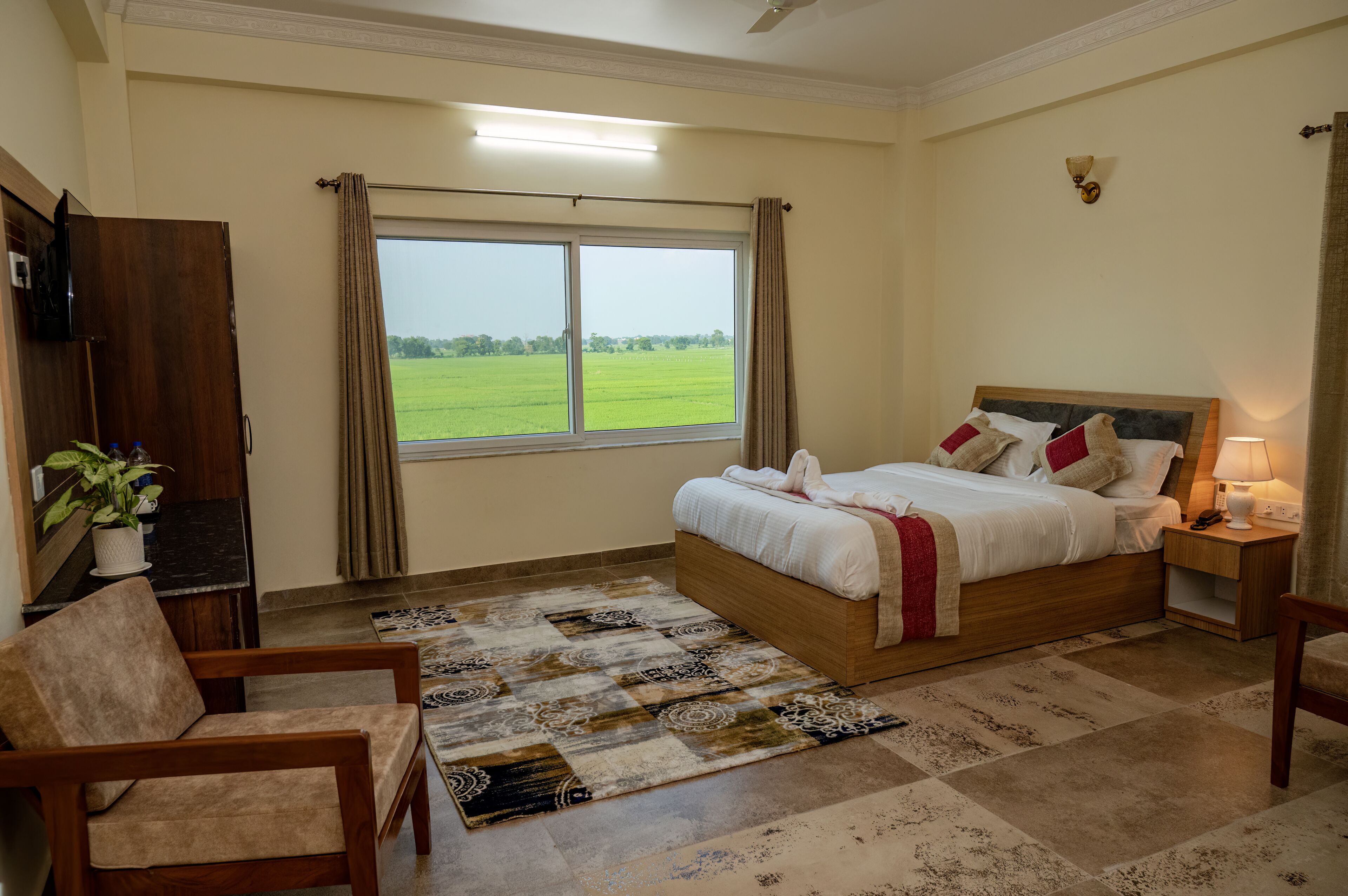 A bedroom with a large bed, a chair, a desk, a window with a view of a green field, and a ceiling fan.