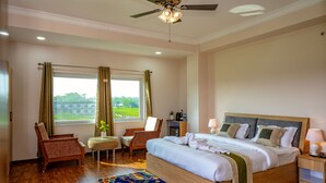 A bedroom with a large bed, a ceiling fan, a window with curtains, a chair, a small table, and a nightstand. - Lumbini Five Elements Hotel (Lumbini)