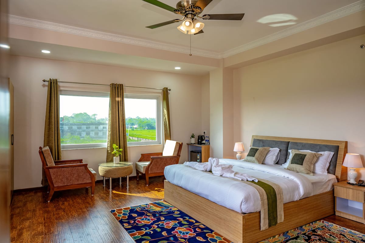 A bedroom with a large bed, a ceiling fan, a window with curtains, a chair, a small table, and a nightstand.