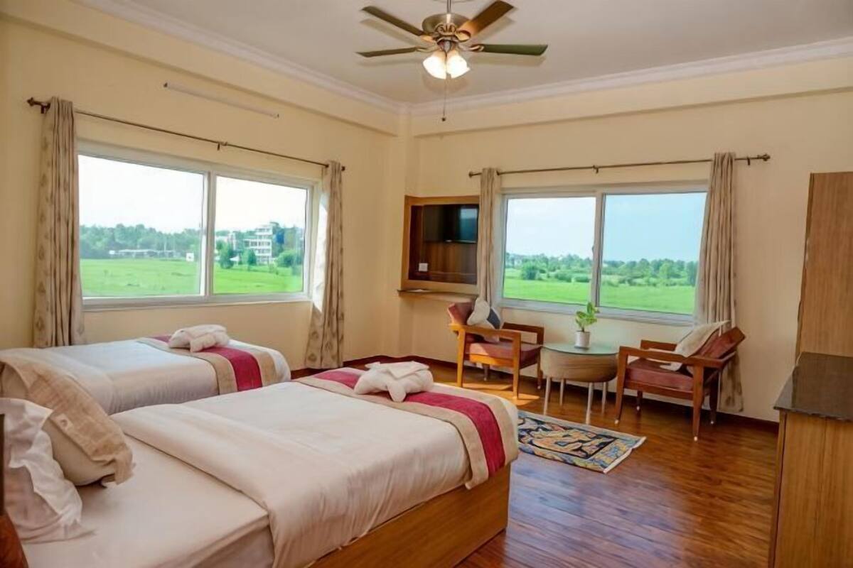 A hotel room with two beds, a desk, a chair, a television, and large windows with a view of greenery.