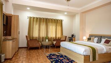 Quarto de hotel com cama, escrivaninha, duas cadeiras, uma pequena mesa com uma planta e um ventilador de teto.