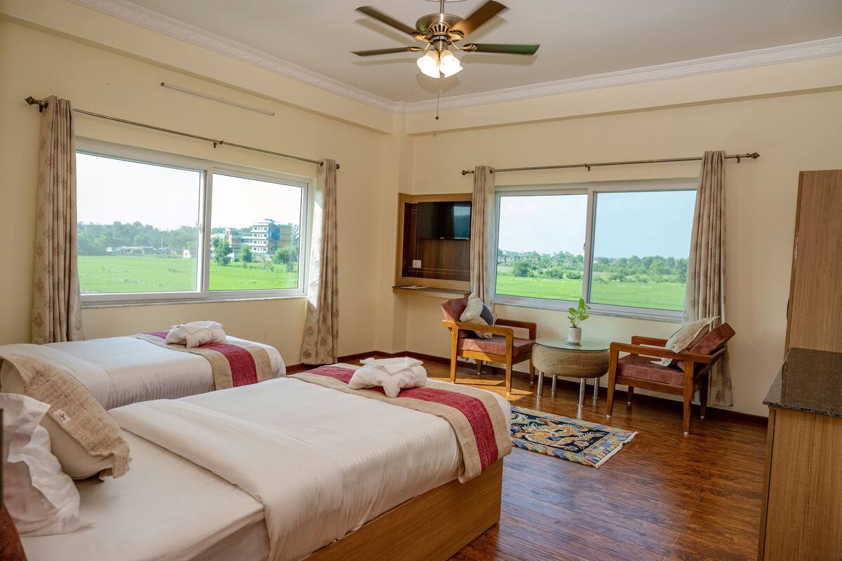 A hotel room with two beds, a ceiling fan, a TV, a small table with a plant, and large windows with curtains.