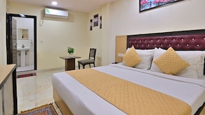A hotel room with a bed, a desk, a chair, a small table with a plant, and a bathroom visible through an open door. - Hotel Hari International (Noida)