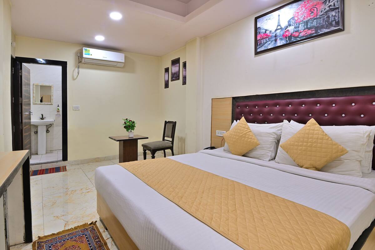 A hotel room with a bed, a desk, a chair, a small table with a plant, and a bathroom visible through an open door.