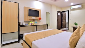 A hotel room with a bed, a desk with a flat-screen TV, a wardrobe, and an air conditioning unit. - Hotel Hari International (Noida)