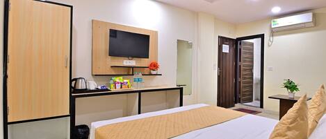 A hotel room with a bed, a desk with a flat-screen TV, a wardrobe, and an air conditioning unit.