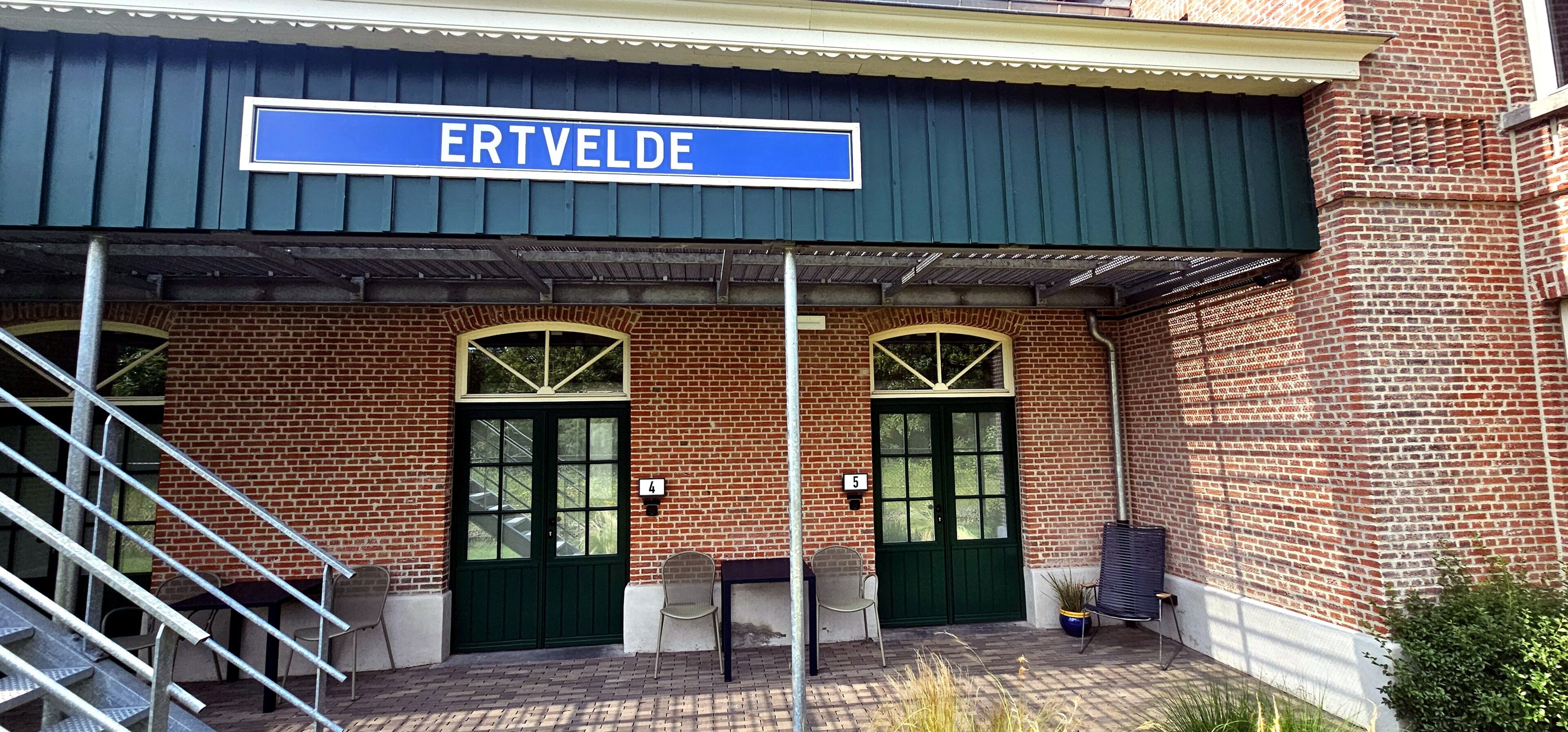 A brick building with a sign reading 'ERTVELDE.' There are two green doors, a covered entrance, and outdoor seating with chairs and tables.