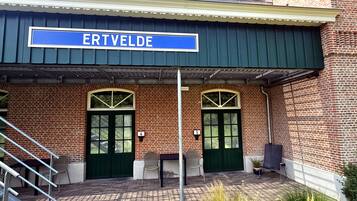 A brick building with a sign reading 'ERTVELDE.' There are two green doors, a covered entrance, and outdoor seating with chairs and tables.