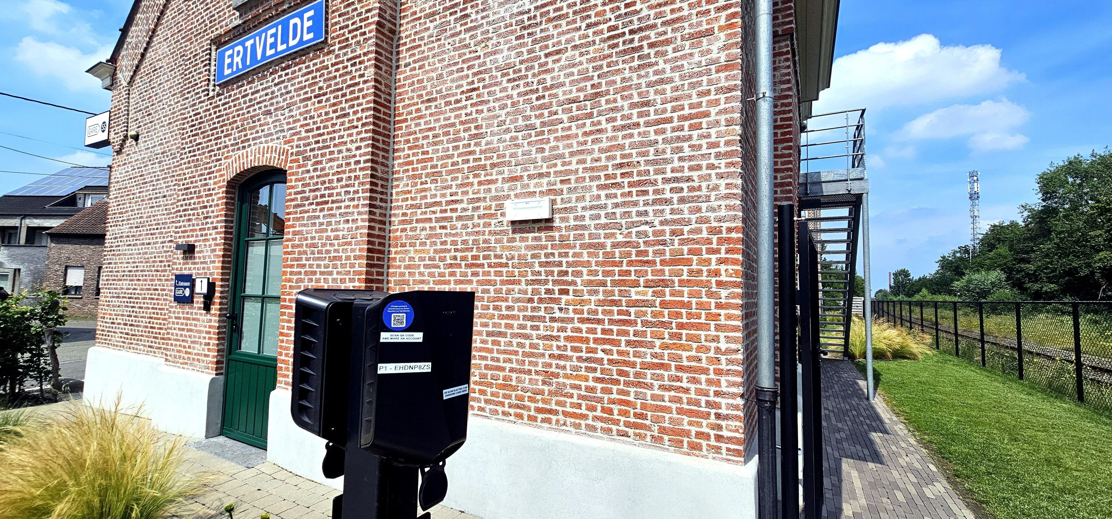 Un bâtiment en briques sur lequel on peut lire l’inscription « ERTVELDE », et une boîte noire portant un code QR.