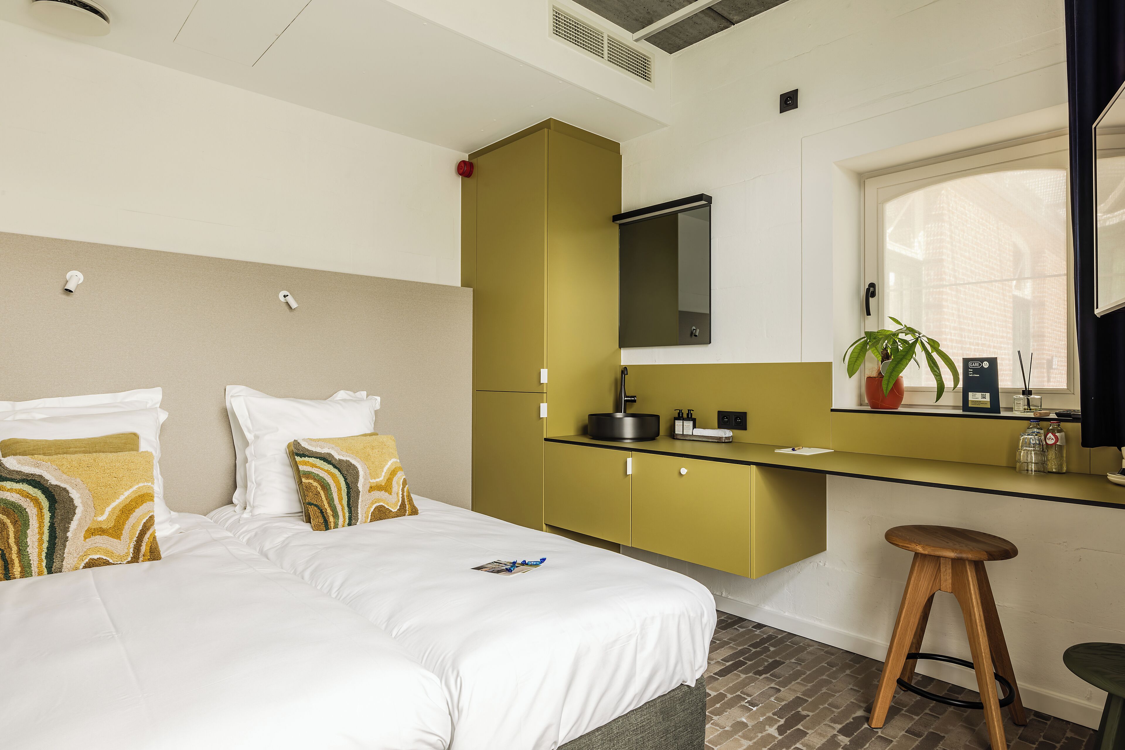 A hotel room with a bed, a small kitchenette, a window with a view, and a wooden stool.