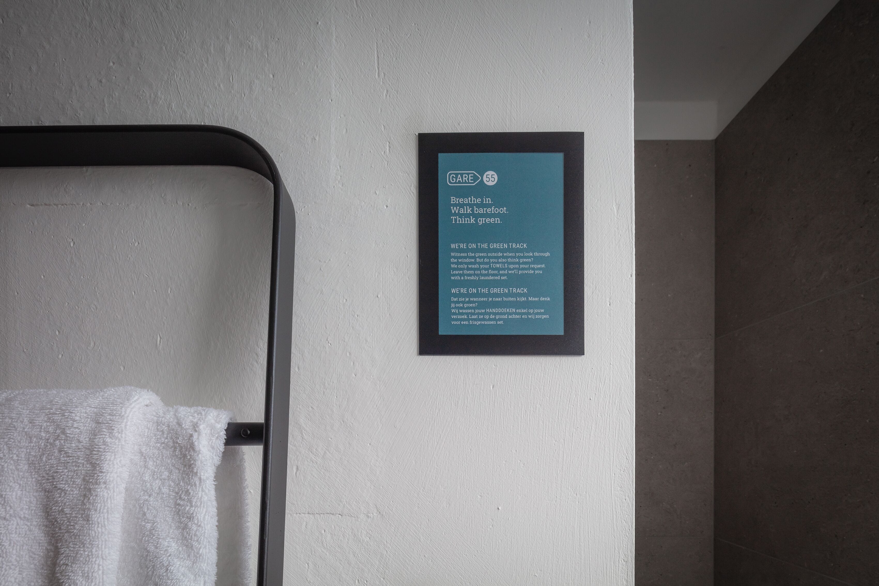 A framed sign with text on a wall, a towel hanging on a rack, and a corner of a room with a dark floor.