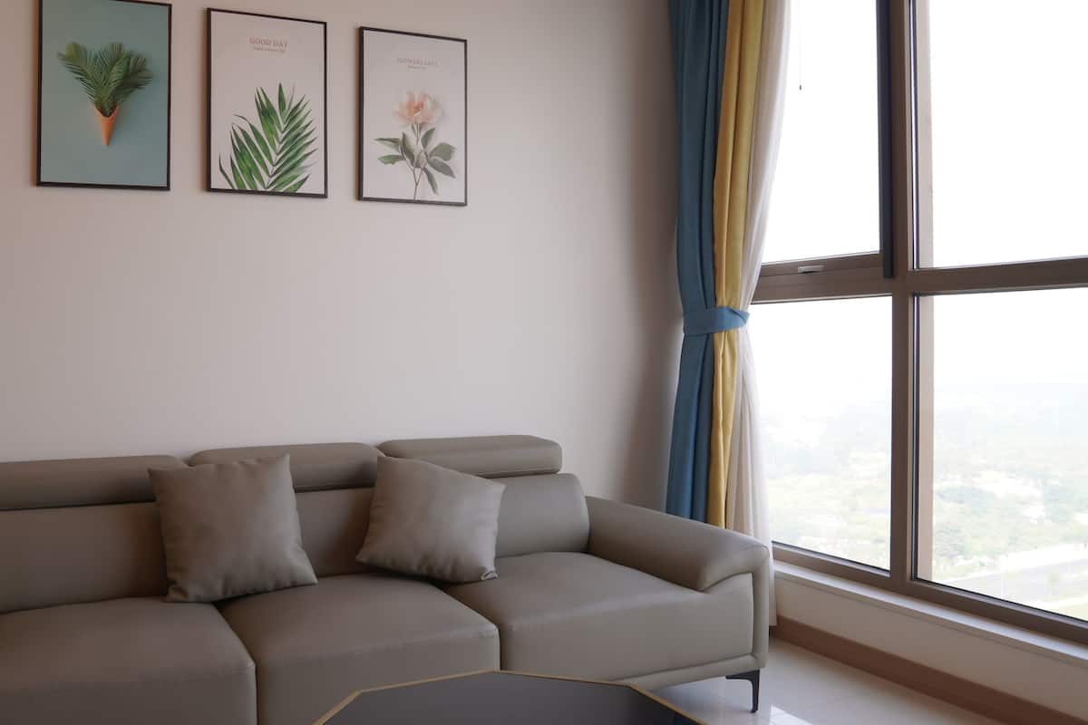 Una sala moderna con un sofá seccional gris, tres láminas botánicas enmarcadas en la pared y un ventanal con cortinas.