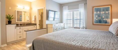 Een slaapkamer met een groot bed, een kaptafel met spiegel, een televisie en een raam met jaloezieën.