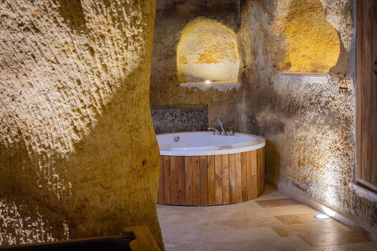A rustic bathroom with a wooden bathtub, stone walls, and a small shelf with a light fixture.