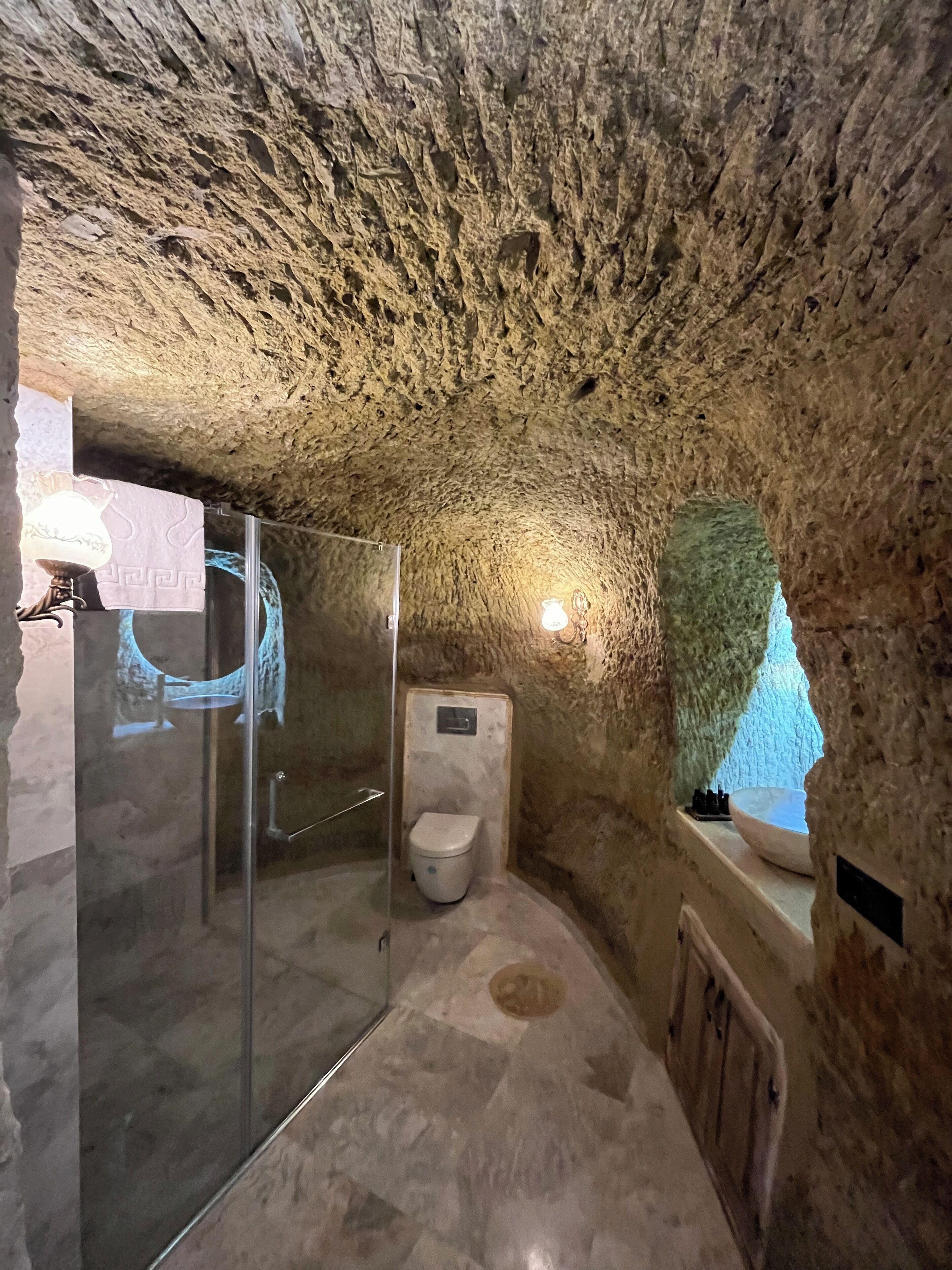 Un baño con techo de piedra, cabina de ducha de vidrio, inodoro y lavamanos con ventana superior.