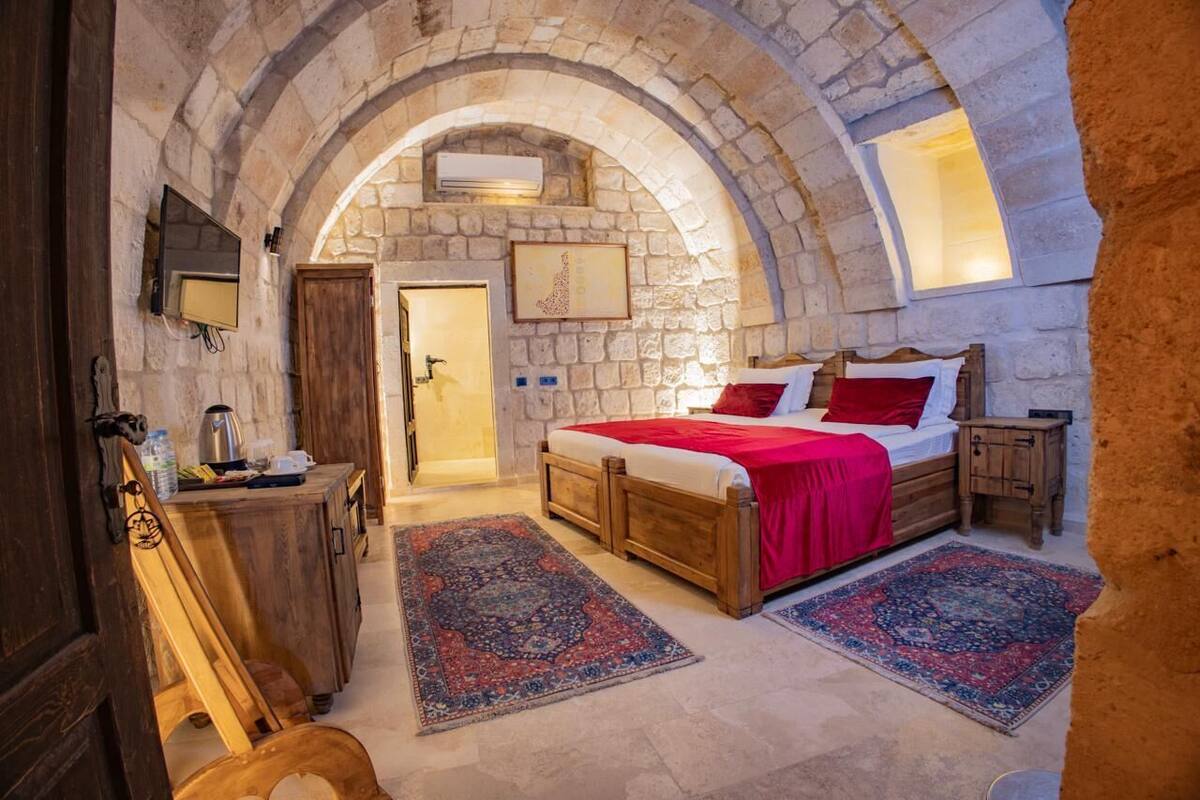A bedroom with a stone archway, wooden bed, red bedding, and two rugs.
