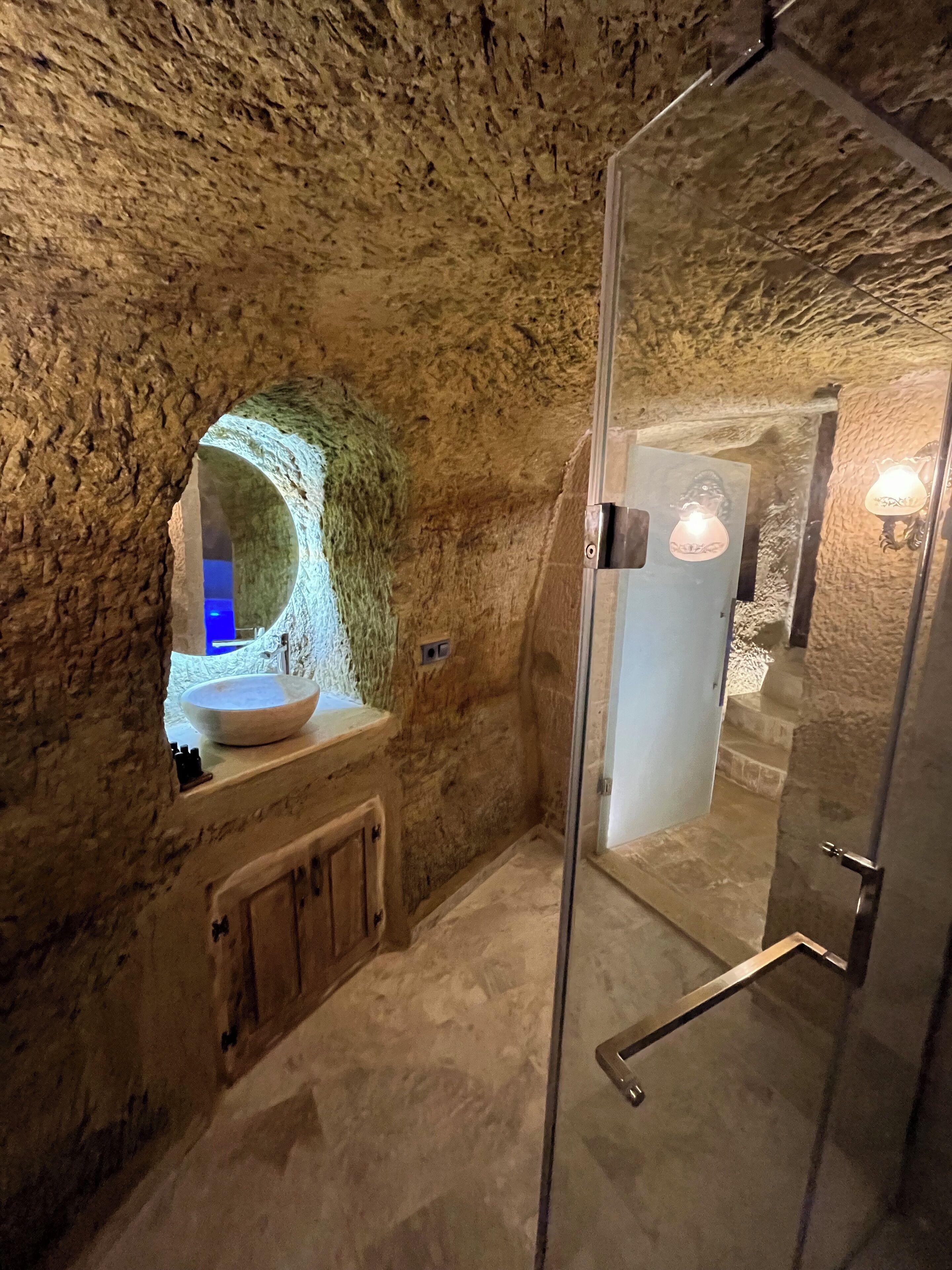 Un baño con paredes de piedra, una ducha con cerramiento de vidrio, un lavamanos redondo y un mueble de madera debajo.
