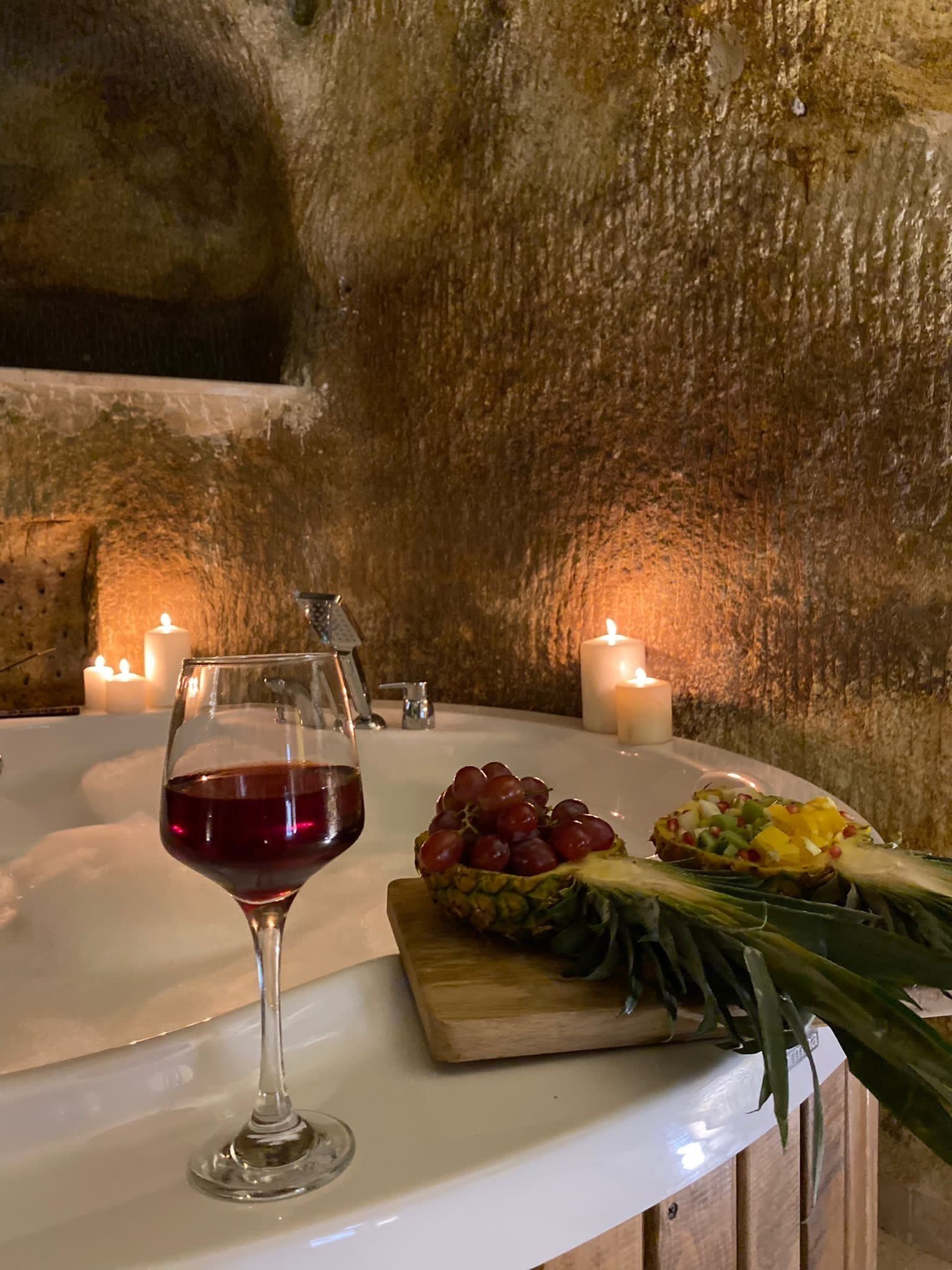 A table with a wine glass, grapes, and a pineapple on a white countertop, surrounded by candles.