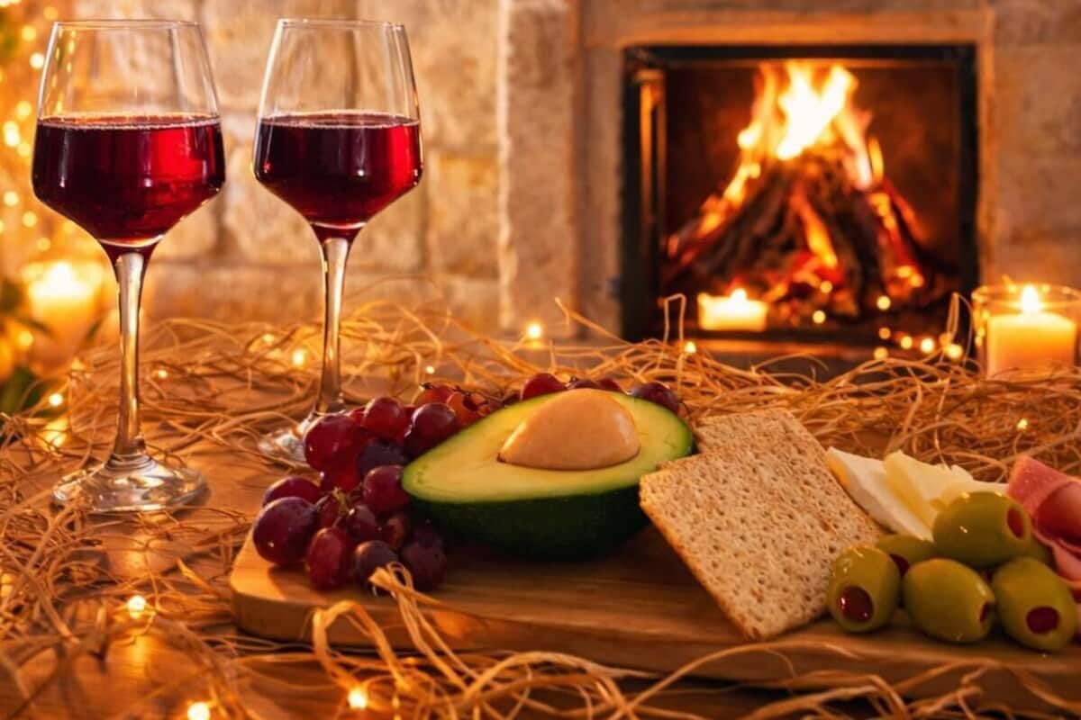 A cozy room with a fireplace, two glasses of red wine, an avocado, grapes, olives, and bread on a wooden table.