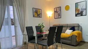 Un salon comprenant une table à manger en verre, quatre chaises, un canapé et une lampe. Trois œuvres d’art encadrées ornent le mur, et une fenêtre est agrémentée de rideaux.