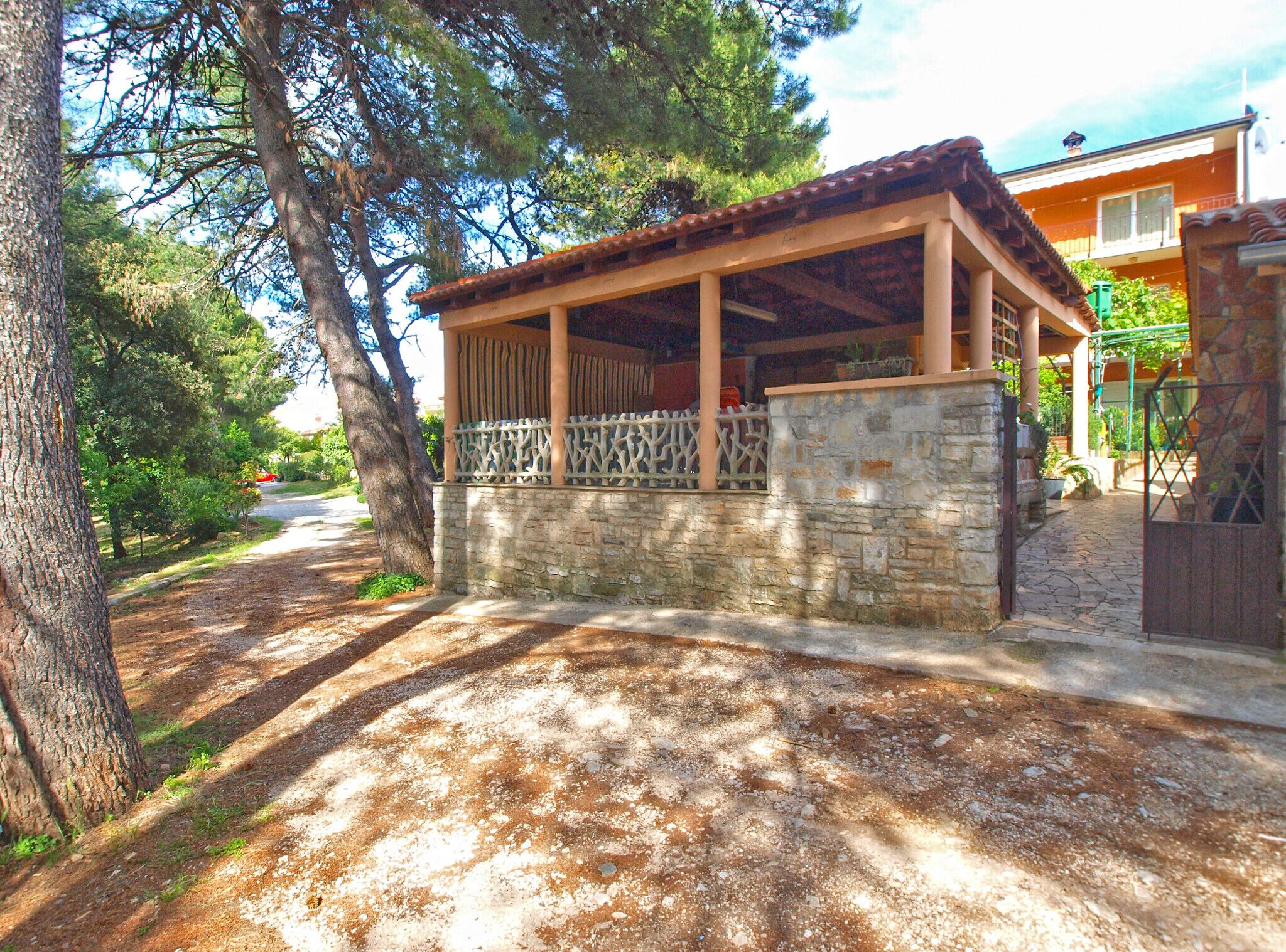 A covered outdoor area with stone walls and a wooden roof, surrounded by trees and a paved path.