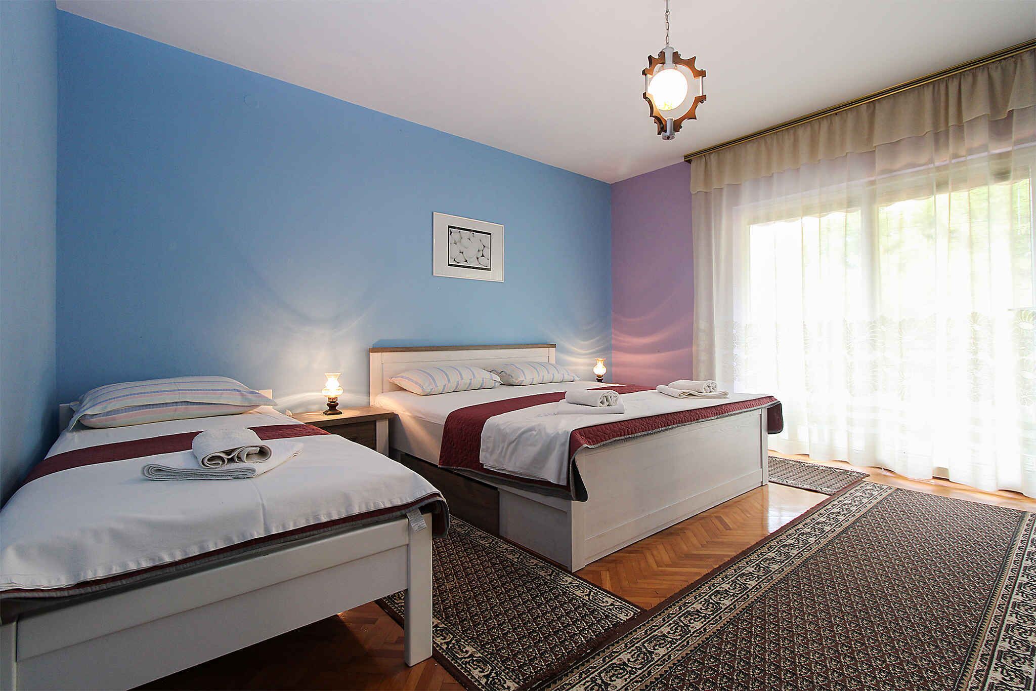 A double room with two beds, a large window with sheer curtains, a hanging light fixture, and a patterned rug.