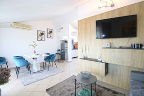A modern living room with a dining area, a flat-screen TV mounted on a wooden wall, and a kitchen in the background. - Holiday apartment with air conditioning (Pula)
