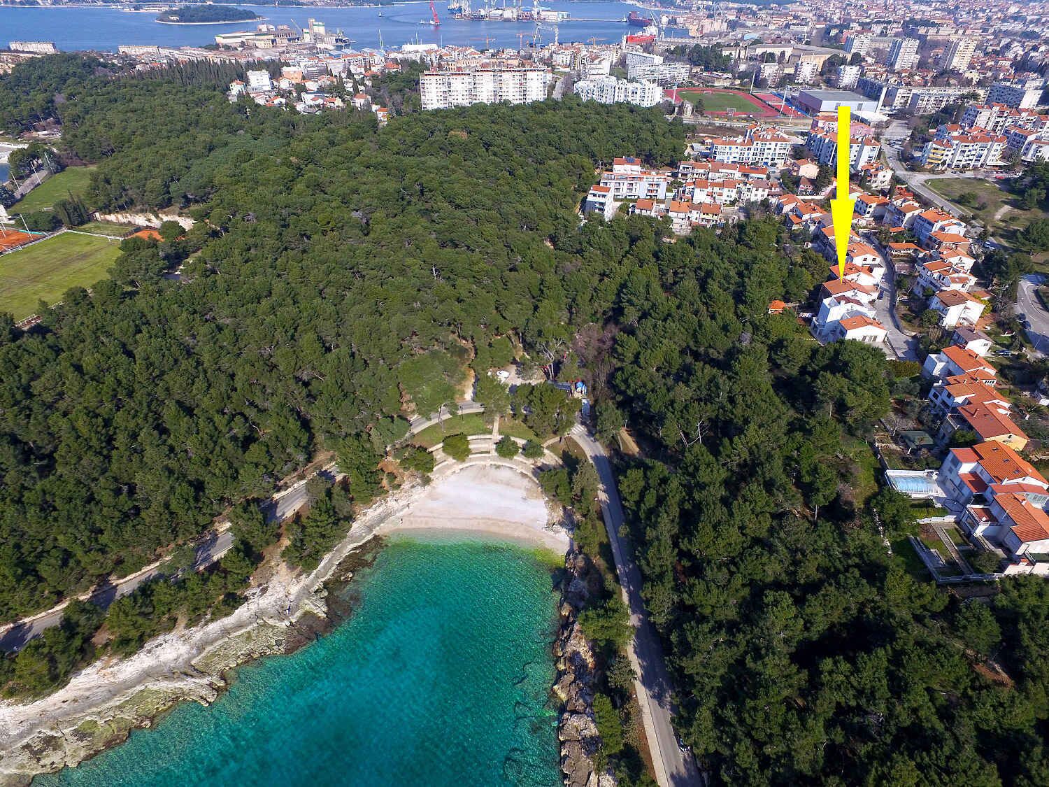 An aerial view of a coastal area with a beach, dense forest, and residential buildings.