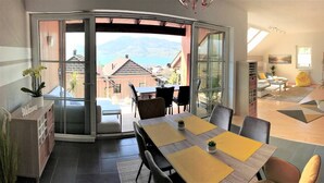 A modern dining area with a view of a mountain and a lake, a sliding glass door leading to an outdoor seating area, and a cozy living space with a sofa and a coffee table. - Apartment With Amazing Lake- and Mountain View (Kaisigen)