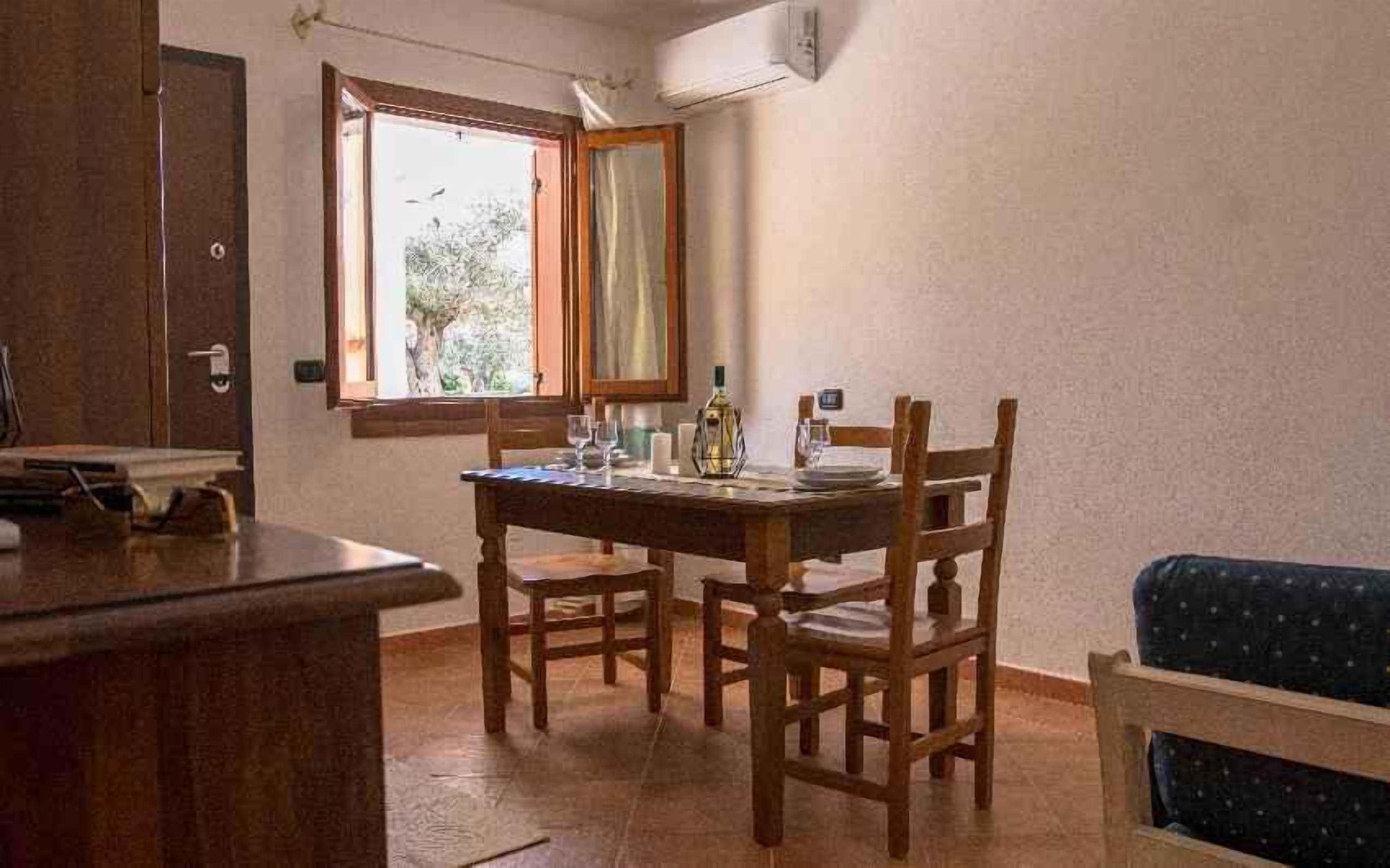 A dining area with a wooden table and chairs, a window with a view of greenery, and a door to the left.