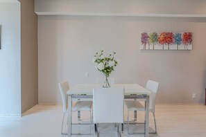 Un coin repas avec une table et des chaises blanches, un vase rempli de fleurs et un tableau coloré représentant un arbre accroché au mur.