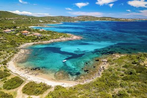 Vue aérienne d’une zone côtière avec des eaux turquoise limpides, une plage de sable et quelques bâtiments dispersés.
