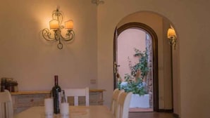 Uma sala de jantar com uma mesa comprida, velas e uma garrafa de vinho. Cadeiras rodeiam a mesa e vislumbra-se um jardim através de uma porta em arco.
