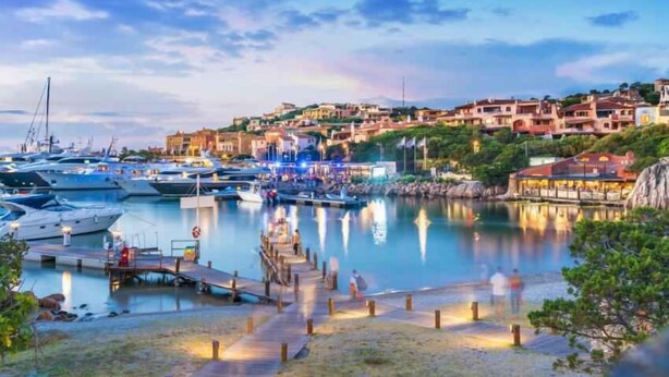 Uma marina com barcos atracados, um calçadão à beira-mar e edifícios ao fundo.