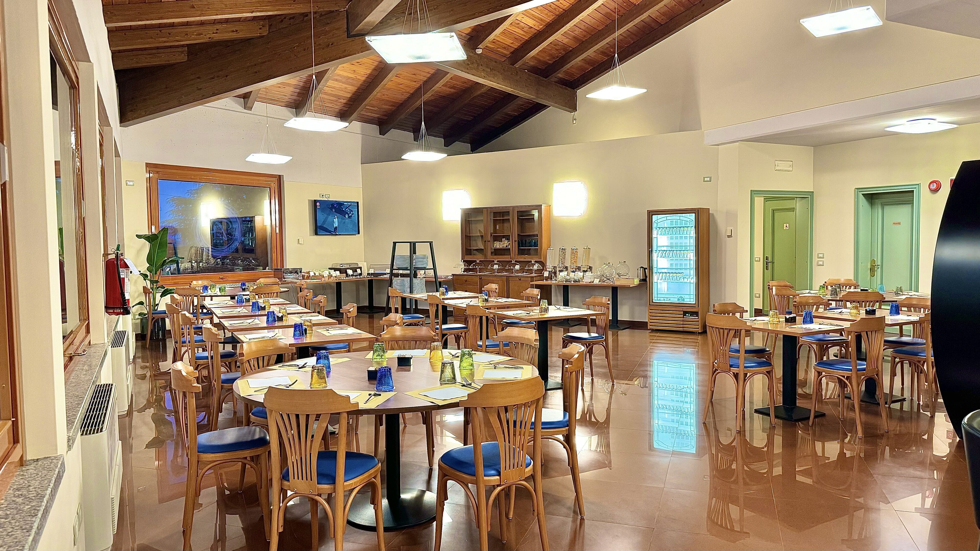 A spacious dining area with wooden tables and chairs, a large window with a view, and a green door in the background.