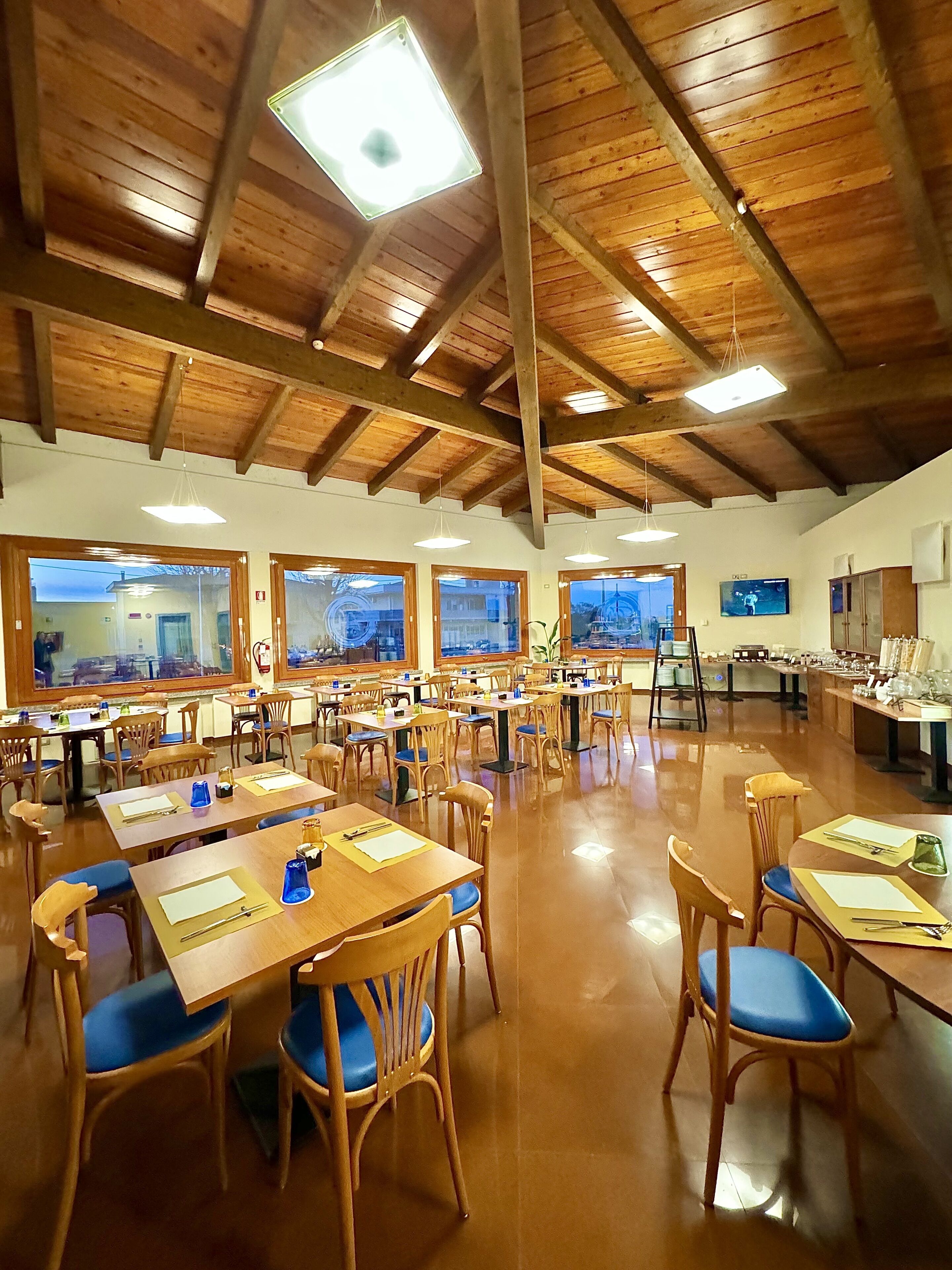 Un vaste espace dédié à la salle à manger, avec des tables et des chaises en bois, de grandes fenêtres et un plafond haut avec des poutres apparentes.