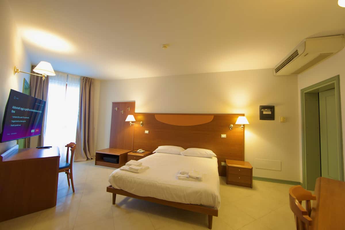 A hotel room with a bed, bedside tables, a desk with a chair, a television, and a door.