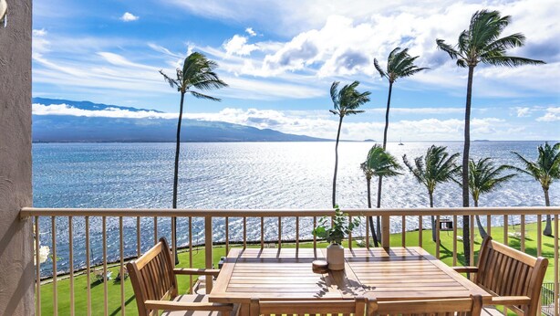 A wooden deck with a table and chairs, overlooking a body of water with palm trees and a distant mountain range.