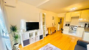 Ein kompakter Wohnraum mit Flachbildfernseher an einer weißen Unterhaltungsanlage, einem Essbereich mit Holztisch und -stühlen, einer Küche mit weißen Schränken und einem Herd sowie einem großen Fenster mit Vorhängen.