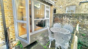 Eine Terrasse mit Glastür zu einem Raum, weiße Gartenmöbel und eine Ziegelwand.