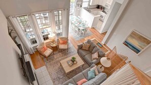A spacious living area with a gray sofa, a coffee table, and a dining table set for six. A kitchen with white cabinets and a dining area with a view through large windows is visible.