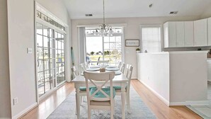 A dining area with a table set for four, white chairs, a chandelier, and a view through large windows to the outdoors.