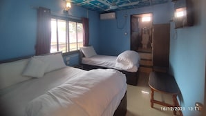 Una habitación con dos camas, un ventilador de techo, una ventana con cortinas, una puerta que da a un baño y una pequeña silla de madera.