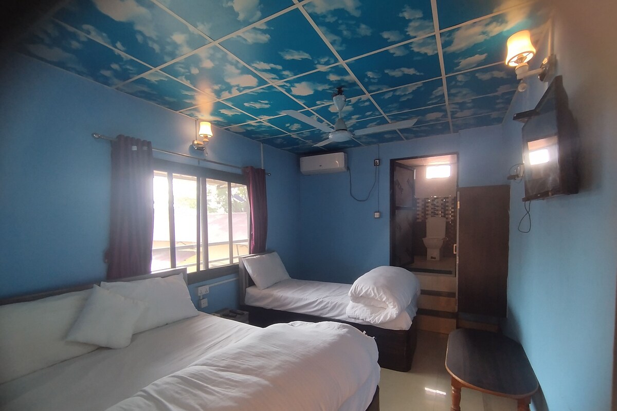 Una habitación con dos camas, un ventilador de techo, una ventana con cortinas, una puerta que da a un baño y una pequeña silla de madera.