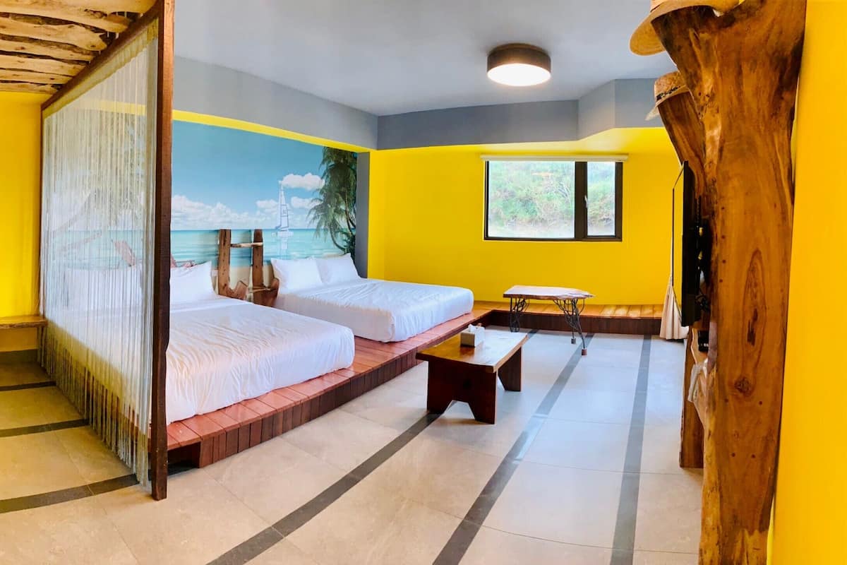 A room with two beds, a bench, a small table, and a mural of a beach scene.