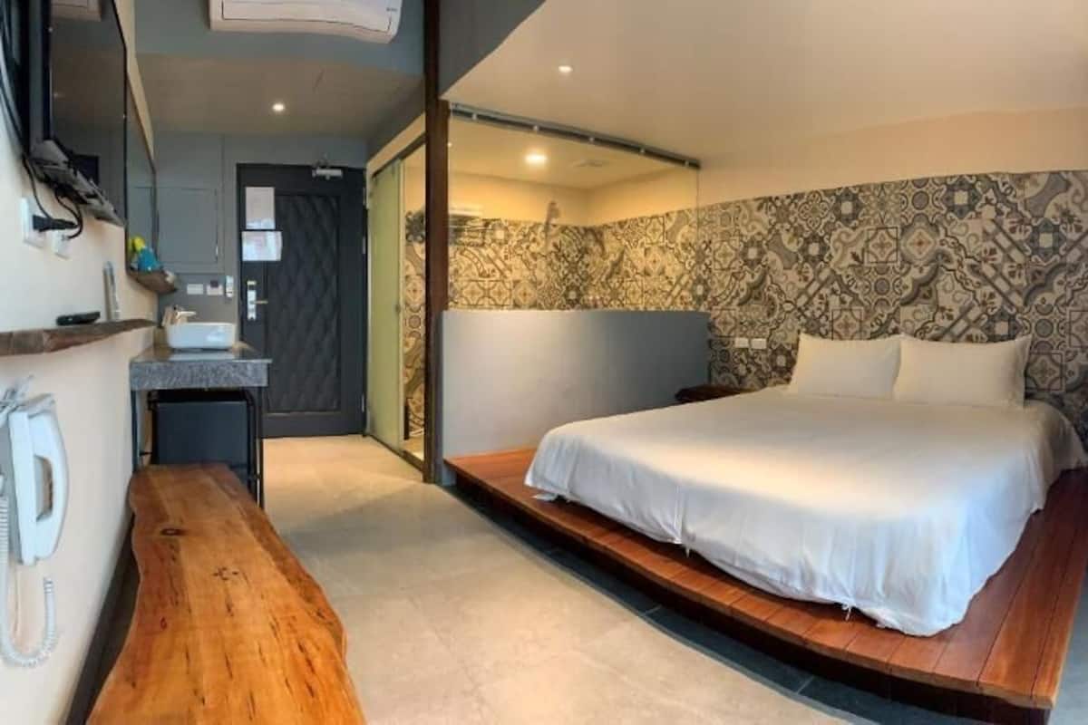 A modern hotel room with a large bed, a wooden bench, a built-in desk, and a bathroom with a mirror and sink.